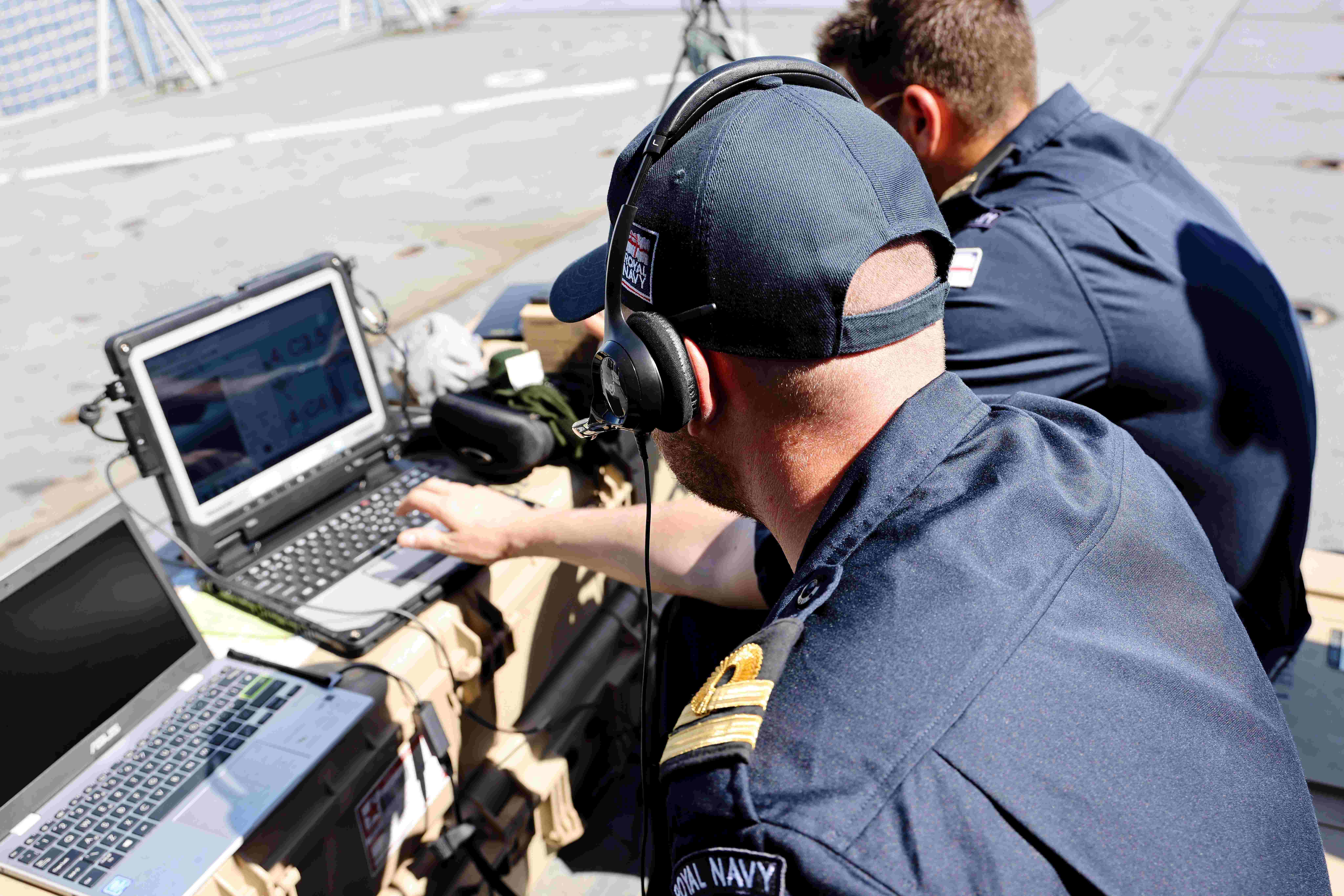Two Royal Navy Officers on deck of ship, sat at computers with headsets on and in blue uniforms